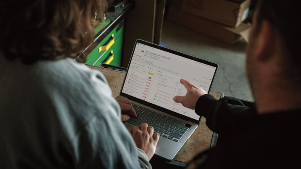 Two people review John Deere’s Operations Center PRO Service on a laptop, showing diagnostic trouble codes and repair tools for equipment maintenance.