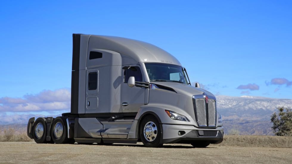 A Kenworth T680 tractor-trailer with a 76-inch sleeper cab parked on a dirt lot with mountains in the background under a clear blue sky.