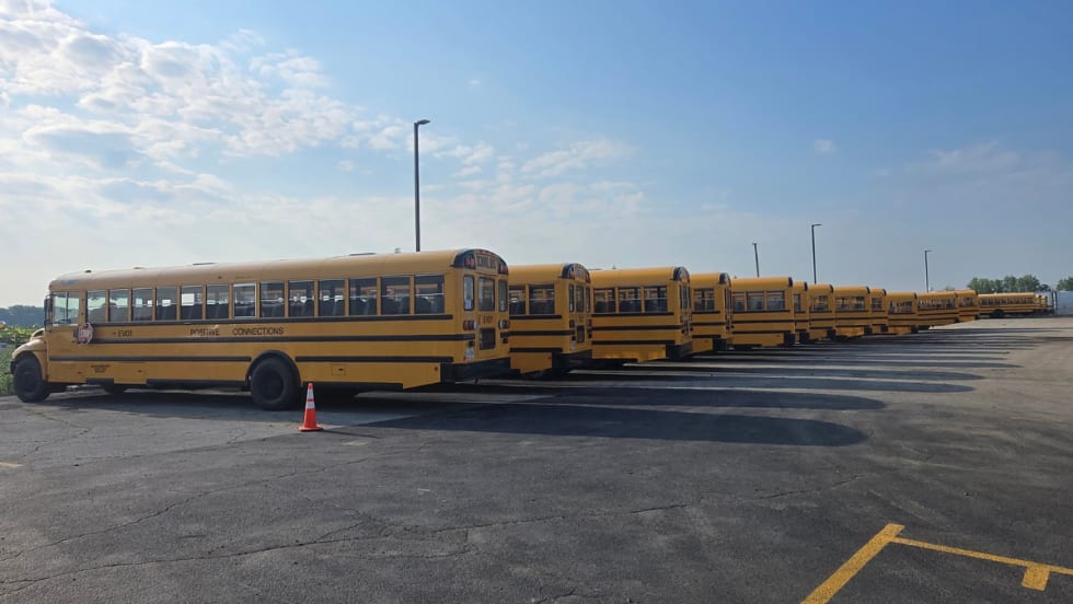 Row of yellow International CE Series electric school buses parked in a lot in Bloom Township, Illinois, ready for deployment. One bus is marked “EV01” with an orange cone nearby.
