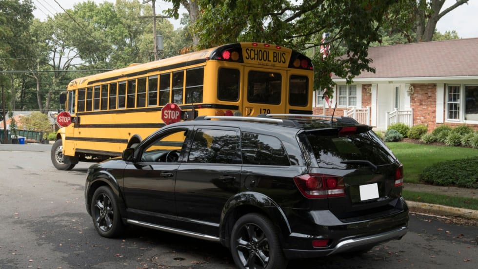 A black SUV stops behind a yellow school bus with its stop arms extended and red lights flashing in a residential neighborhood, indicating children may be boarding or exiting the bus.