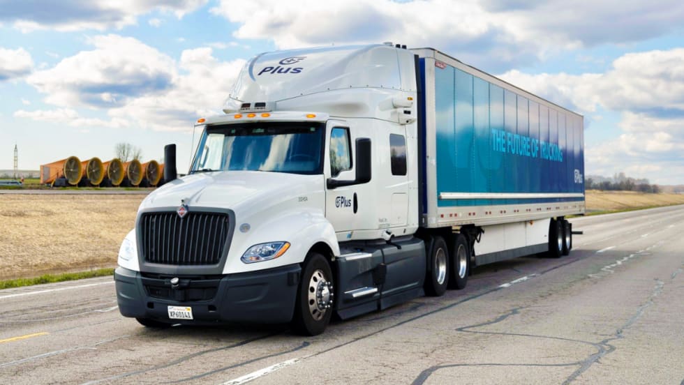 A white Plus autonomous semi-truck with a blue trailer reading "The Future of Trucking" drives on an open road under a partly cloudy sky, promoting self-driving freight technology.