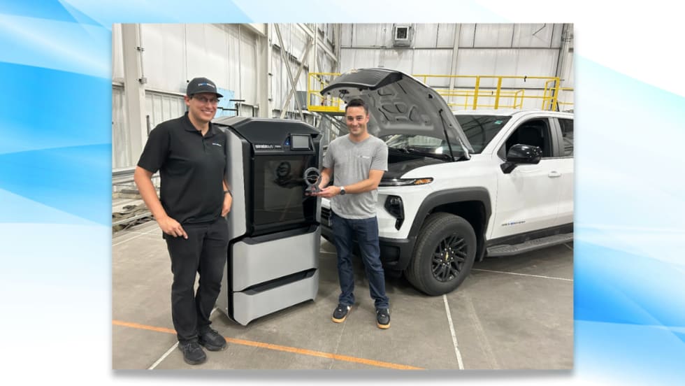 Two GM engineers stand beside a 3D printer holding a trophy, with a white Chevrolet Silverado EV Max Range Work Truck in the background and hood open after a record-setting range test.