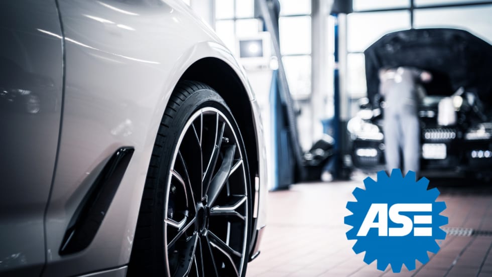 Close-up of a car wheel in an auto repair shop with a mechanic working in the background; ASE logo displayed in the foreground.