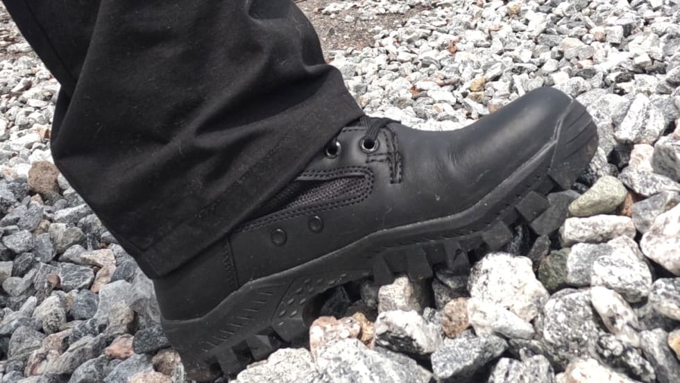 Black tactical boot with rugged sole standing on gray stones and gravel