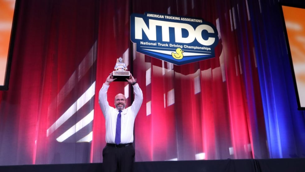 David Comings holds the NTDC Grand Champion trophy above his head on stage at the 2025 ATA National Truck Driving Championships.