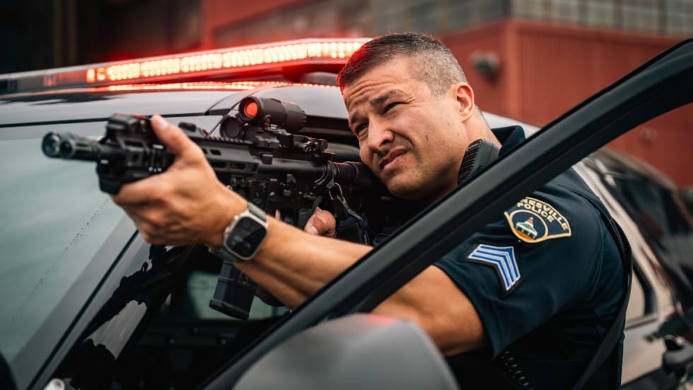 Police officer aiming tactical rifle from patrol vehicle with emergency lights