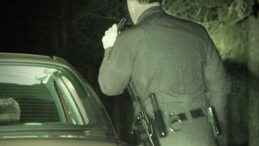 Police officer at night standing beside patrol car, viewed from behind