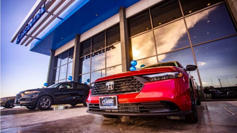 Exterior of the new Classic Honda of Midland dealership with floor-to-ceiling glass, blue Honda entry frame, and vehicles on display, including a red Honda sedan in the foreground.