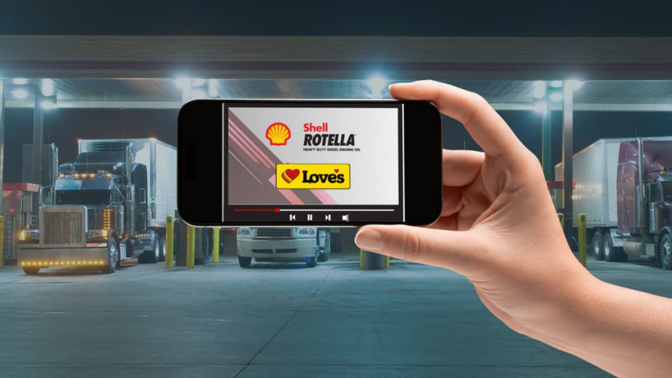 Hand holding a smartphone displaying a Shell Rotella and Love’s Travel Stops video screen, with semi-trucks parked at a brightly lit fuel station in the background.