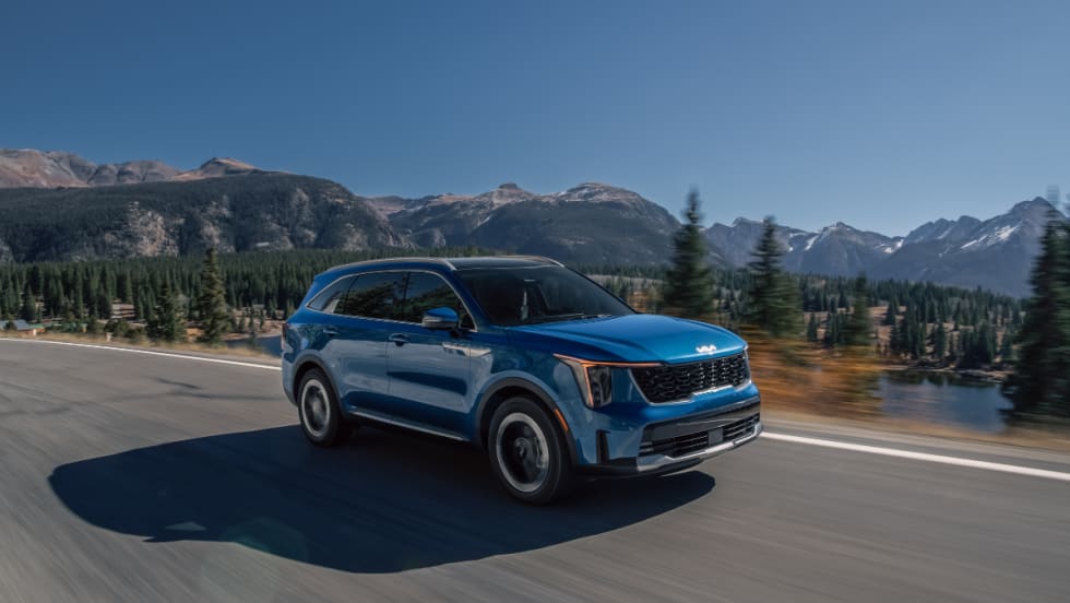 Blue 2026 Kia Sorento Hybrid driving on a mountain highway with trees and water in the background.