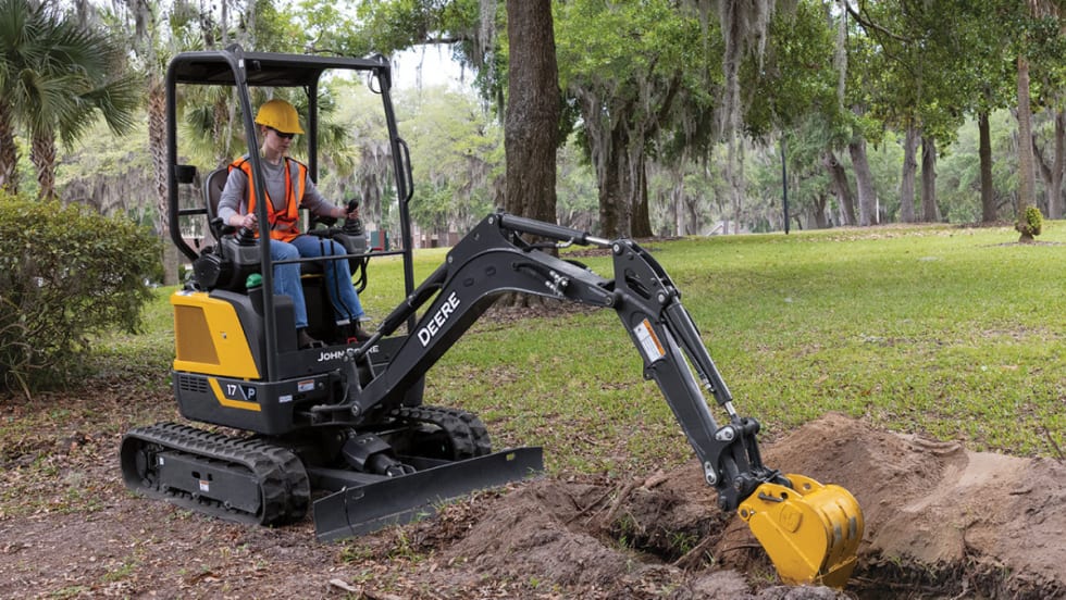 Operator using a John Deere 17 P-Tier compact excavator to dig in a grassy park area surrounded by trees.
