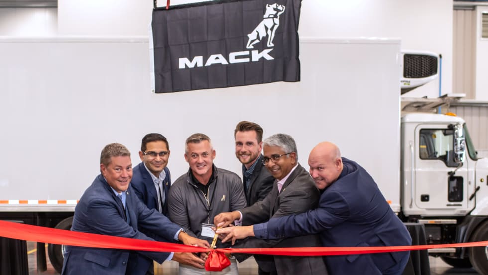 Group of businessmen cut ribbon in front of a white box truck with a Mack Trucks sign on the side. 