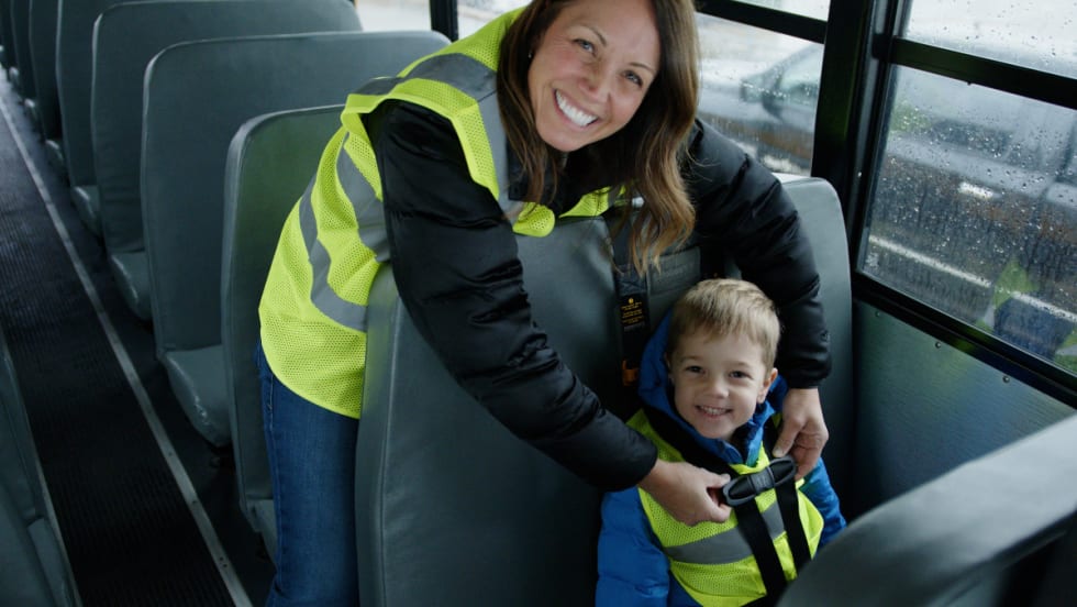 A school bus driver helps a young student buckle up while smiling inside a bus, reflecting First Student’s First Serves behavioral support program.