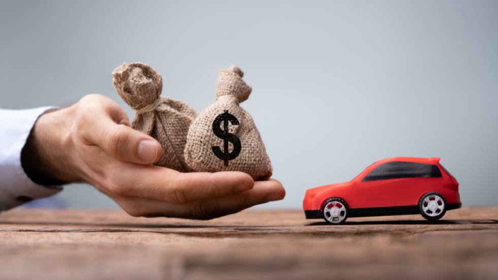 A hand holding small burlap money bags next to a toy red car, symbolizing auto financing, loan payments, and dealership profitability.