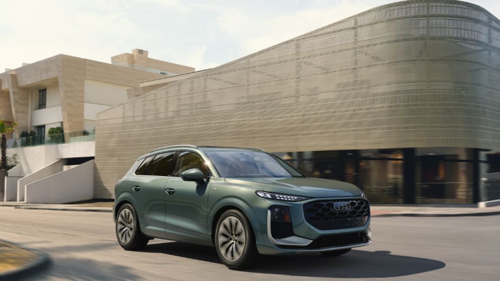Green 2026 Audi Q3 compact SUV driving on a city street past a modern building in daylight.