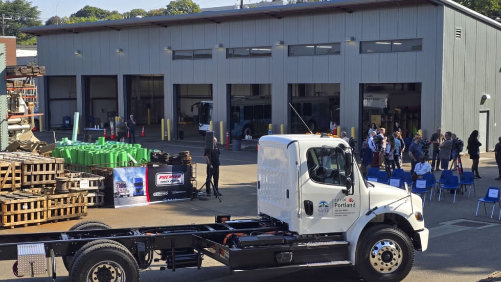 White City of Portland Freightliner eM2 electric truck parked in front of a maintenance building with event seating and attendees in the background.