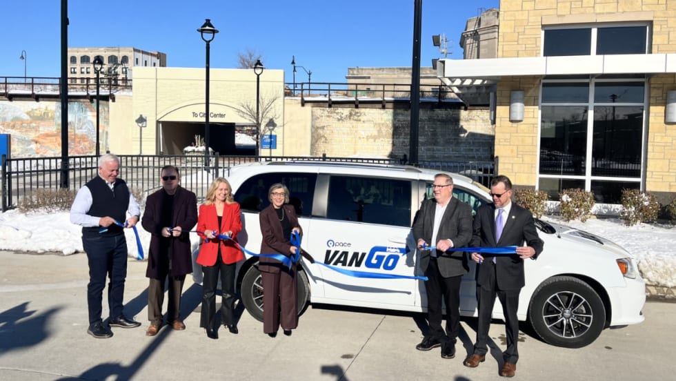 A group of officials stands in front of a white Pace VanGo minivan and holds a blue ribbon during a ribbon-cutting event outside a transit facility on a sunny winter day.