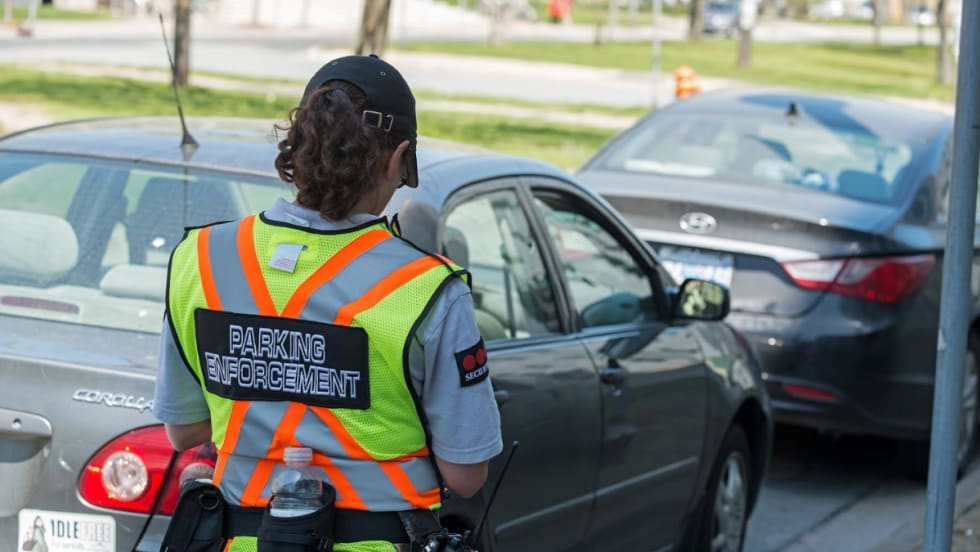 woman officer shown from the back in a vest that states parking enforcement while issuing a parking ticket to a car