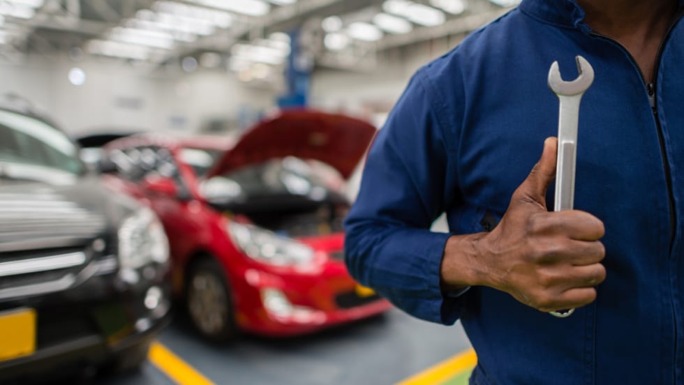 Technician in a blue coverall holding a wrench in an auto repair shop, with cars in the background.