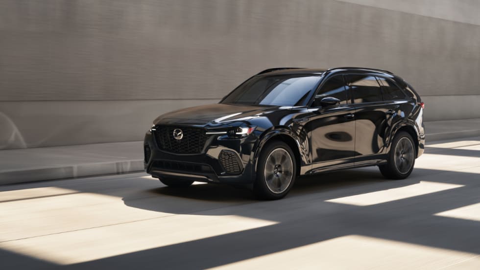 A black Mazda CX-70 crossover driving through an urban roadway with concrete walls in the background.