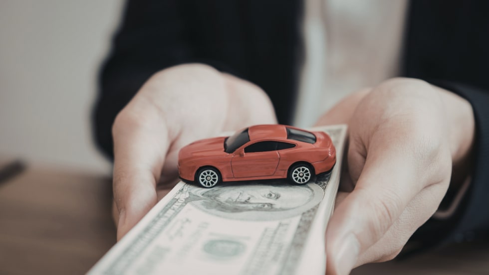 A person holds a stack of cash with a small red toy car on top.