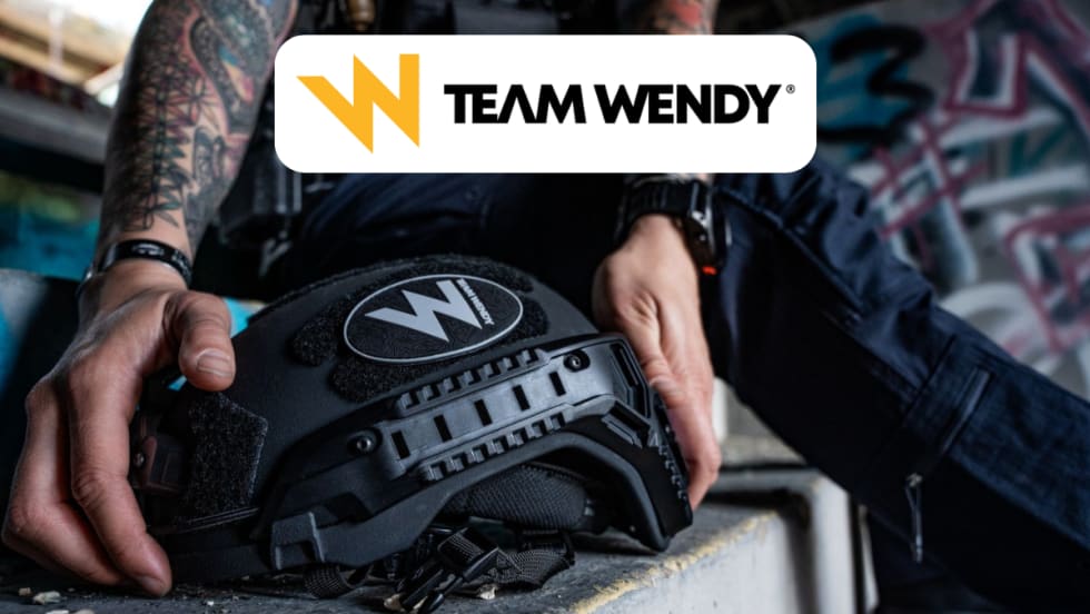 Closeup of hands reaching to pick up a ballistic helmet and a Team Wendy logo in a white box top center in the image.