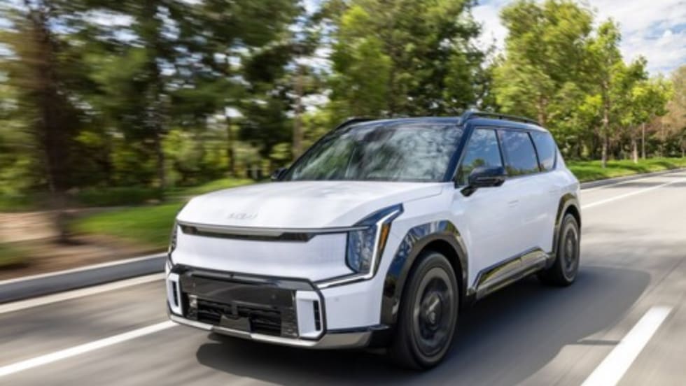 A white 2026 Kia EV9 electric SUV driving on a tree-lined roadway.