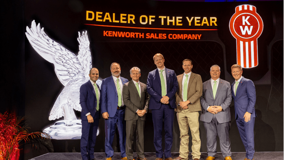 Group of men in business attire on a stage during an award ceremony with background that says Dealer of the Year and a Kenworth logo.