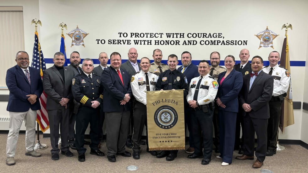 Group of police chiefs in uniform stand in a group at the front of a room following a training class.