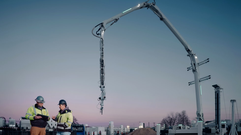 Dark blue sky at dusk, two construction workers, a crane-type structure in the background.