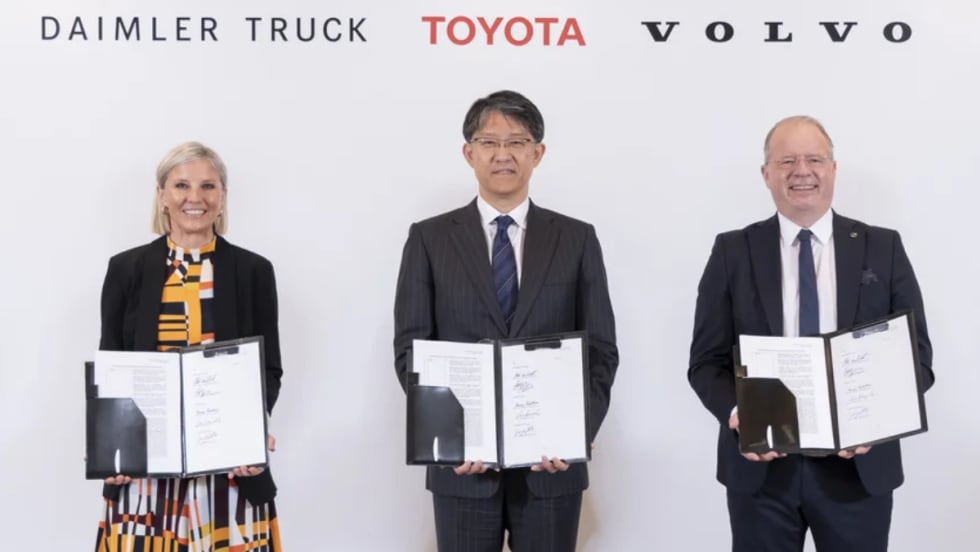 Woman and two men standing holding paperwork agreements they have signed with logos for Daimler Truck, Toyota, and Volvo over their heads.