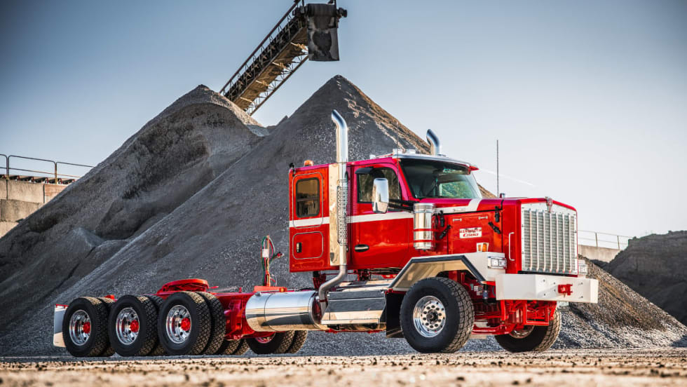 Red Kenworth truck parked sideways with large mound of gravel in background.