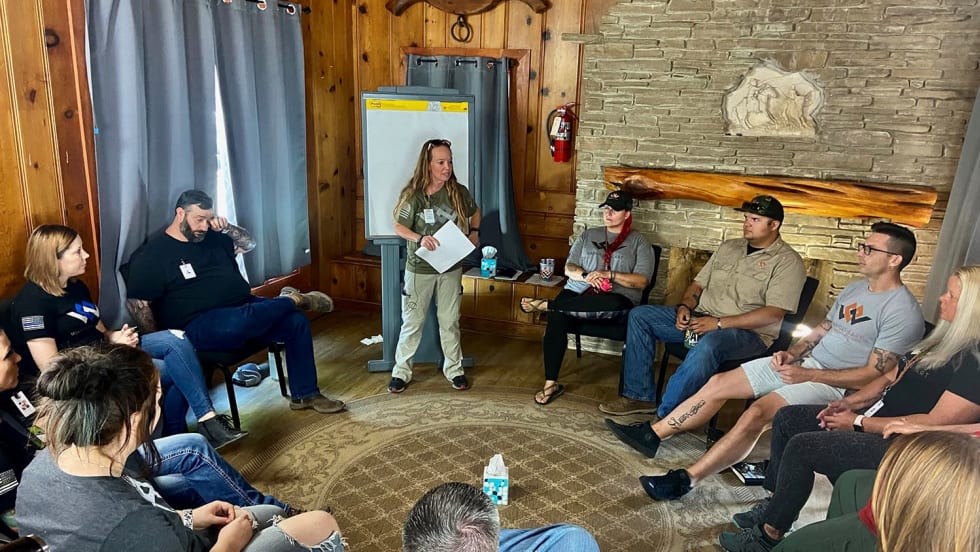 Group of men and women seated in a circle around a room as one woman stands and leads discussion.