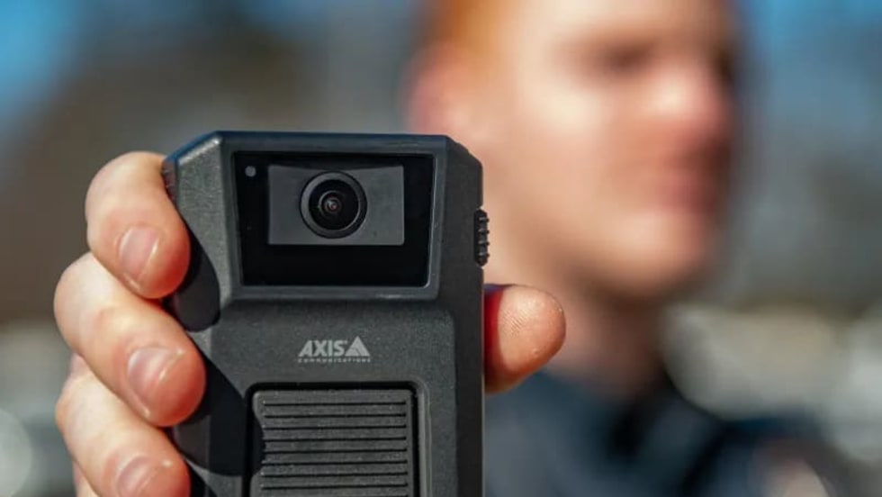 Close-up of a person holding an Axis body-worn camera toward the viewer. The camera is in sharp focus, while the person wearing a dark uniform appears blurred in the background outdoors.