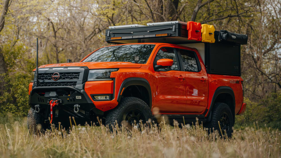 Orange Nissan Frontier PRO-4X overland build parked in a grassy wooded area with off-road tires, heavy-duty front bumper, winch, rooftop cargo storage, and camping accessories mounted above the truck bed.