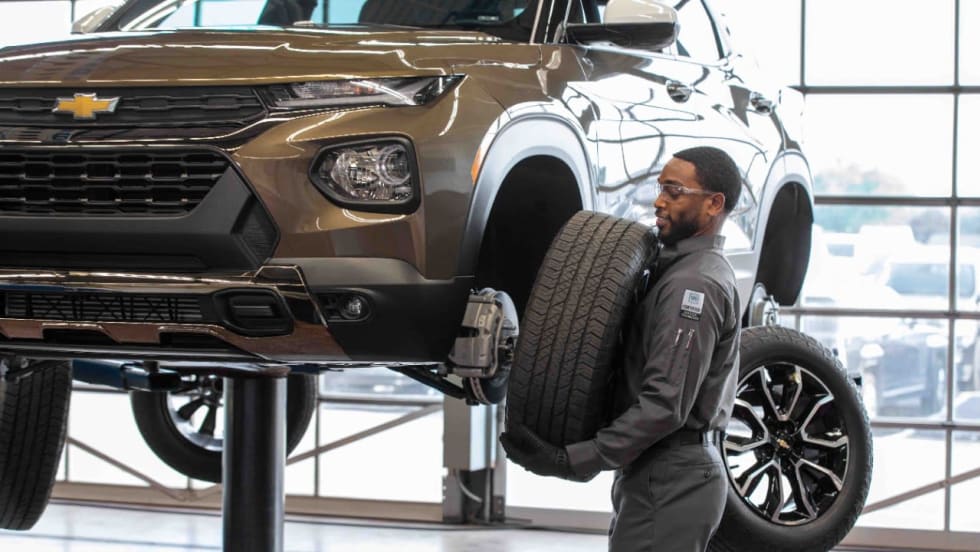 Automotive technician carrying a tire beside a Chevrolet SUV raised on a service lift inside a repair facility. Additional wheels and bright garage windows are visible in the background.