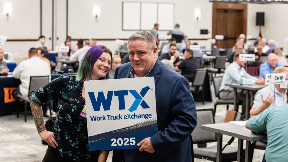Lauren Fletcher and Wayne Parham pose with a Work Truck Exchange 2025 sign during a networking session while fleet professionals meet in the background.