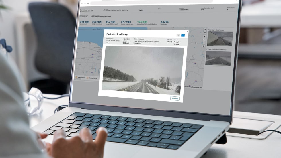 Person using a laptop to view a fleet management dashboard displaying weather alerts, vehicle speed data, maps, and road-condition images. The screen highlights a snowy roadway and a lake-effect snow warning for a fleet vehicle.