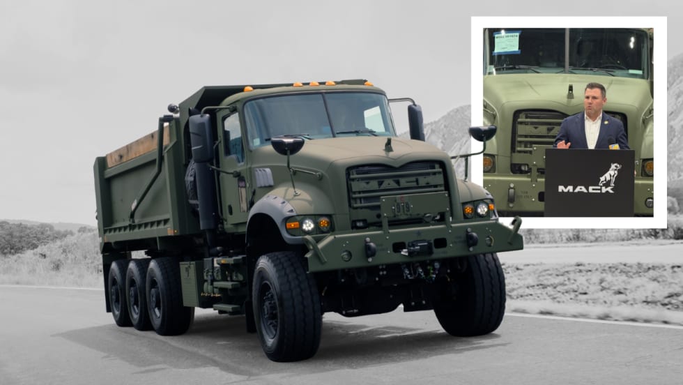 Military-style Mack Defense dump truck parked on a roadway, shown in olive-drab paint with rugged off-road features. An inset photo shows a speaker at a Mack-branded podium in front of the truck during a presentation or announcement.