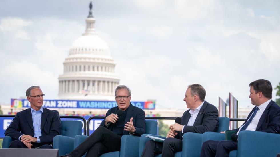 Four men in suits on the National Mall with giant video screen showing capitol building in the background