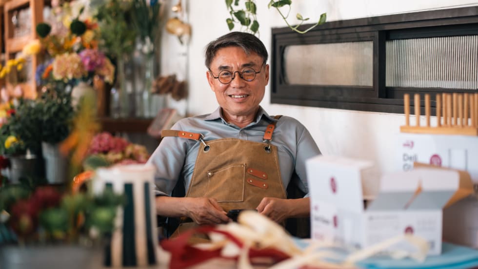 Flower shop owner wearing glasses and an apron sits behind a worktable surrounded by flowers, packaging supplies, and small boxes.