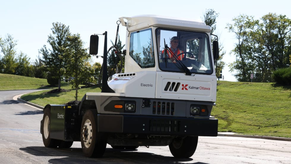 Kalmar Ottawa electric terminal tractor drives on a paved roadway with an operator in the cab, showcasing the zero-emission yard truck in an outdoor commercial fleet setting.