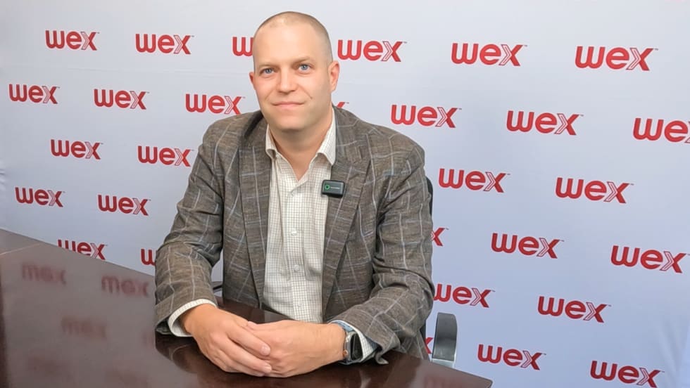 Business professional seated at a conference table in front of a WEX-branded backdrop, wearing a suit jacket and lapel microphone, during an interview or executive discussion about fleet payment and fuel management solutions.