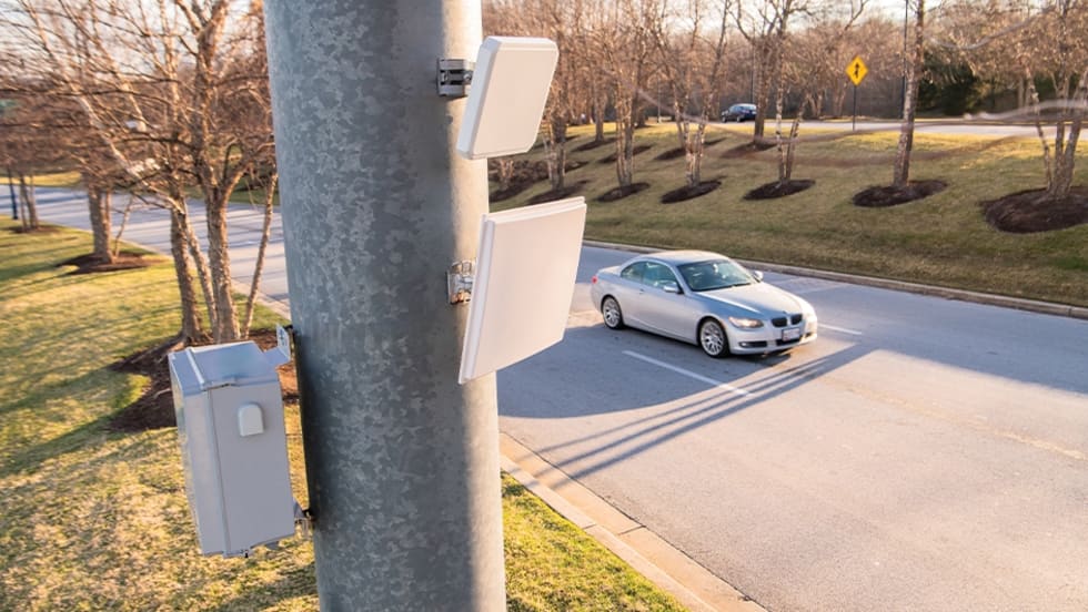 Roadside traffic monitoring equipment mounted on a utility pole overlooks a passing car, illustrating wireless vehicle detection technology used for traffic management, signal optimization, or roadway data collection.