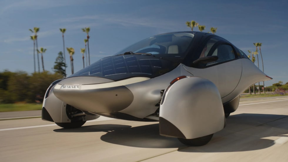 Futuristic silver three-wheeled Aptera solar electric vehicle drives along a palm-lined road, featuring integrated solar panels, aerodynamic bodywork, enclosed front wheels, and a streamlined teardrop-shaped cabin.
