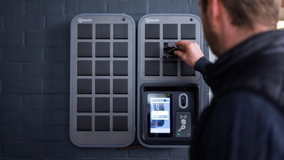 A man removes a vehicle key from a Keycafe electronic key cabinet mounted on a wall.