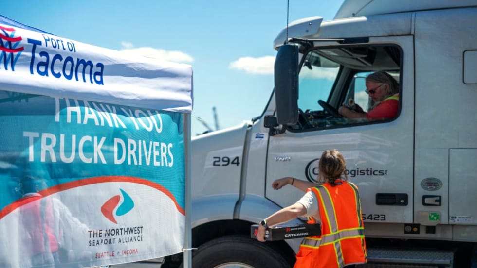 Port of Tacoma truck driver appreciation event, with a worker in a safety vest handing food to a semi-truck driver beside a “Thank You Truck Drivers” banner.