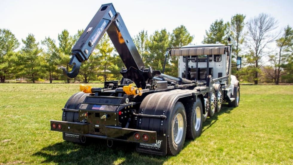 Rear three-quarter view of a heavy-duty truck equipped with a raised hydraulic hooklift system, parked on grass with trees in the background.