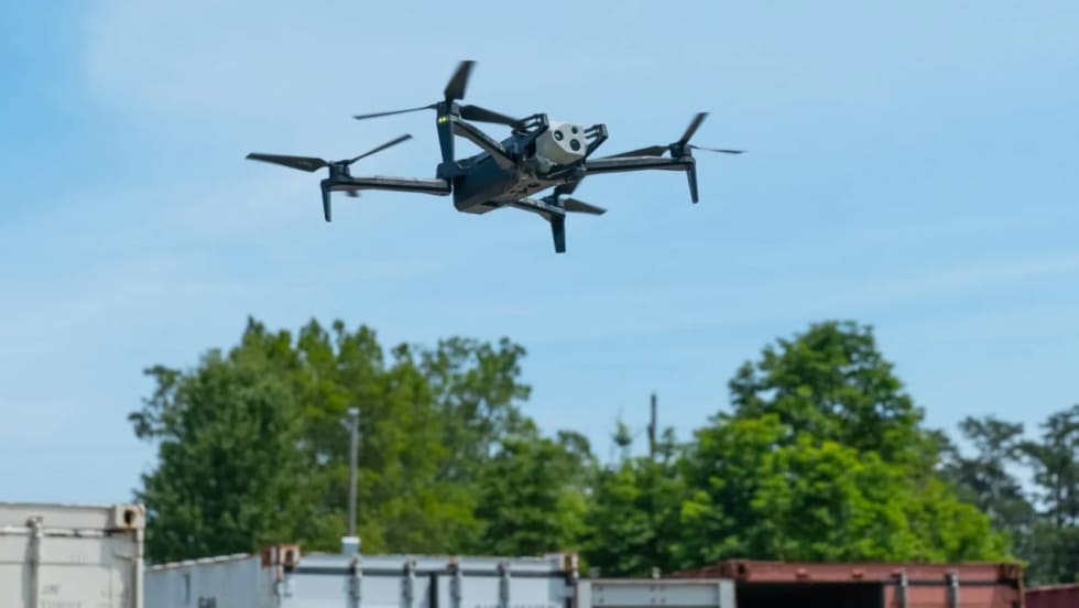 A HAWK drone flies above an industrial area with trees, buildings, and shipping containers visible below.