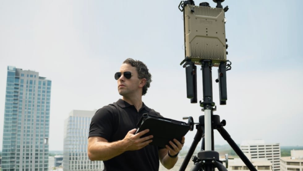 A technician wearing sunglasses holds a tablet beside tripod-mounted electronic detection equipment on a rooftop, with office buildings visible in the background.